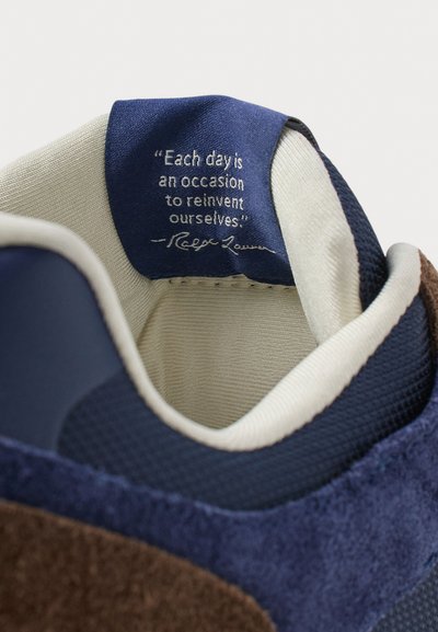 Adidași navy și maro cu un design din suede texturat și plasă. În interior, o etichetă albastră afișează un citat motivant în font alb.
