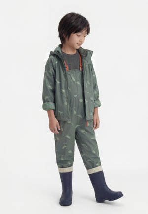 Dziecko stojące, ubrane w zieloną kurtkę przeciwdeszczową z nadrukiem dinozaura oraz ogrodniczki, w ciemnych gumowych butach, patrzące w dół na bok.