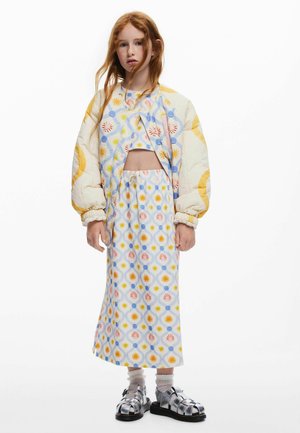 Fetiță tânără cu păr lung roșcat, purtând o jachetă matlasată cu imprimeu floral pastel, top scurt și fustă asortate, șosete albe cu volane și sandale argintii.