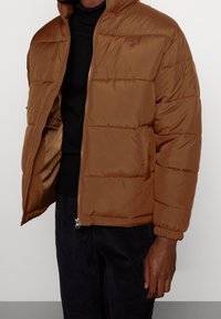 Brązowa kurtka puchowa z zamkiem z przodu, poziomym przeszyciem i kieszeniami po bokach. Tkanina wydaje się gładka, z miękką teksturą i kapturem.