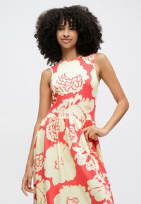 Žena s kučeravými vlasmi, ktorá nosí červené šaty bez rukávov s veľkými krémovými kvetinovými vzormi, pózuje s jednou rukou na boku na jednoduchom pozadí.