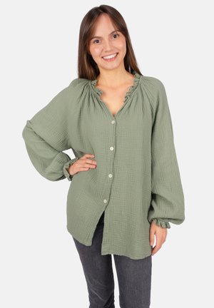 Jeune femme souriante, portant un chemisier texturé vert clair à boutons avec col et poignets à volants, et un pantalon gris foncé, main sur la hanche.