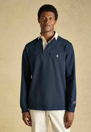 Uomo che indossa una maglia da rugby blu navy a maniche lunghe sopra una camicia bianca con collo, in piedi davanti a uno sfondo beige uniforme.