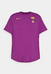 Ljubičasta majica s kratkim rukavima od mekog materijala, s Nike logotipom i FC Barcelona amblemom u žutim i crvenim nijansama na prsima.