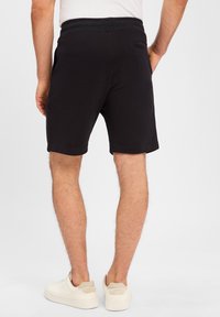 Schwarze Baumwollshorts mit einem eng anliegenden Bund, ausgestattet mit einer Gesäßtasche und einem Saum, der knapp über dem Knie endet, kombiniert mit weißen Turnschuhen.