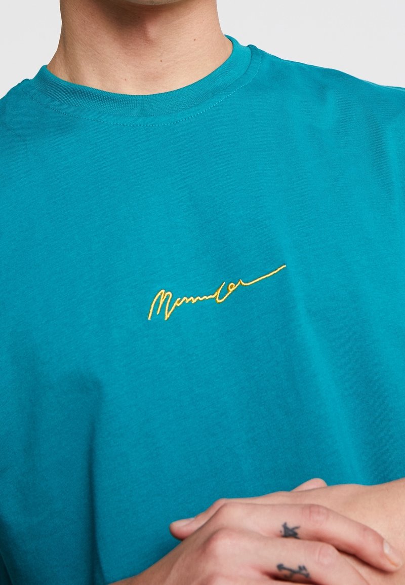 Osoba oblečená v tyrkysovém tričku se žlutým psaným nápisem na hrudi, se zkříženýma rukama, na dvou prstech má malé tetování.