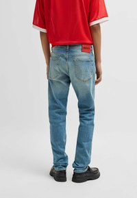 Jeans in denim blu da uomo con vestibilità slim, effetto sbiadito, cinque tasche e una patch rossa con il logo HUGO sul retro della vita. Combinati con una maglietta rossa.