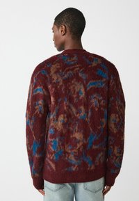 Pull en tricot bordeaux avec une surface texturée et duveteuse, ornée de motifs abstraits bleus et marron, col rond et poignets côtelés.