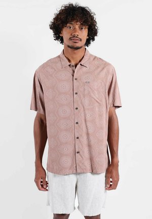 Homme aux cheveux bouclés portant une chemise à manches courtes rose à motifs et un short blanc texturé, debout devant un fond uni.