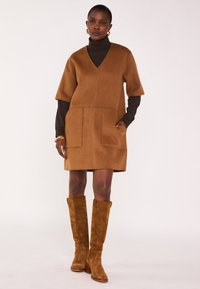 Robe en mélange de laine marron avec col en V, manches courtes, deux poches avant, associée à des bottes hauteur genou en daim marron.