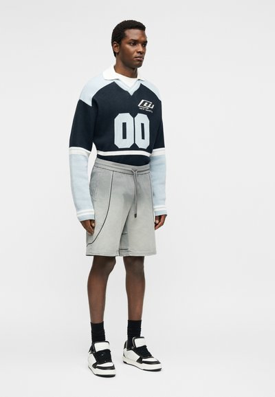 Άνδρας που στέκεται φορώντας μπλε σκούνο και ανοιχτό μπλε πουλόβερ με το "00" και γκρι σορτς, συνδυασμένα με μαύρα και λευκά αθλητικά παπούτσια.