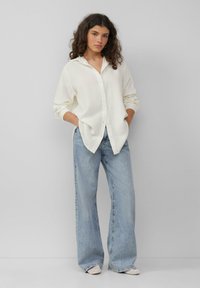 Valkoinen nappikauluspaita, jossa on teksturoitu kangas, yhdistettynä vaaleansinisiin leveälahkeisiin farkkuihin. Malli seisoo kädet puvussa, wear minimalistisia kenkiä.