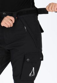 Pantaloni cargo neri in tessuto testurizzato, con vita in Velcro, tasche laterali e un'etichetta con logo. Design resistente con hardware funzionale.