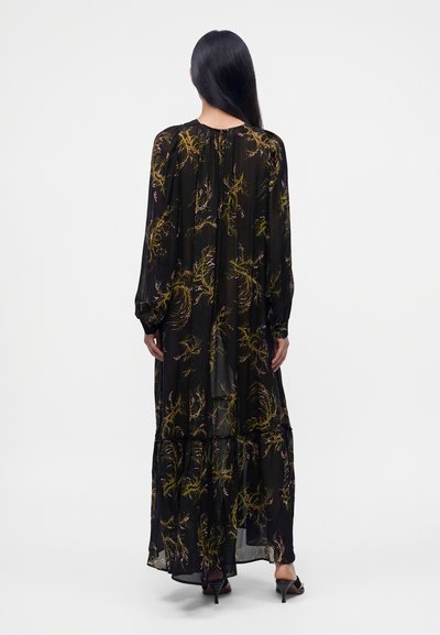 Femme aux longs cheveux noirs porte une robe noire transparente avec des motifs botaniques jaunes et roses et des talons ouverts noirs, debout de dos.