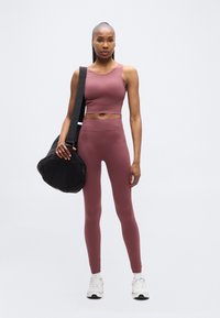 Γυναίκα με μωβ κοντομάνικο τοπ και κολάν, κρατώντας μαύρη τσάντα γυμναστηρίου στον ώμο, στέκεται μπροστά σε απλό λευκό φόντο.