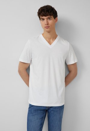 Jonge man met kort krullend haar, gekleed in een effen witte V-hals T-shirt en blauwe spijkerbroek, staand tegen een grijze muur.