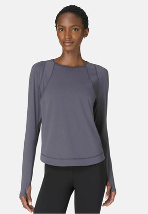 Woman wearing a long-sleeve gray athletic top with thumbholes and black leggings, standing against a plain light background.