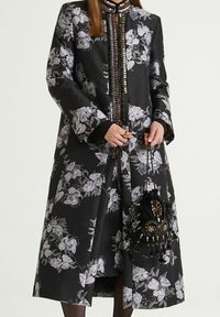 Lungo cappotto nero floreale con tessuto strutturato, caratterizzato da fiori bianchi e argento, collo alto e chiusura frontale dettagliata. Borsa nera con decorazioni.