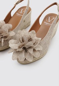 Sandales compensées beiges avec des accents en tissu floral. Elles possèdent un dessus texturé, un dos ouvert et une bride de cheville ajustable. Hauteur d'environ 5 cm.