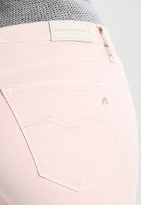 Jeans de mezclilla rosa con una cinturilla texturizada, una etiqueta de cuero con la inscripción "REPLAY JEANS" y un diseño estilizado en el bolsillo trasero.