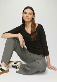 Haut noir à manches longues associé à un pantalon large gris. Baskets blanches avec des accents noirs et une semelle texturée. Modèle assis calmement.