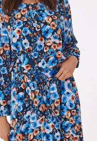 Abito floreale con motivo blu, arancione e nero. Presenta maniche lunghe, collo con bottoni, vita elastica e una cintura per la regolazione.