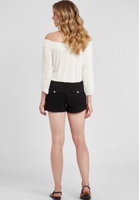 Weißes, schulterfreies Oberteil aus strukturiertem Stoff mit langen Ärmeln, kombiniert mit schwarzen Shorts, die zwei Gesäßtaschen und silberne Knopf-Akzente haben.