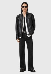 Veste en cuir noir avec deux poches zippées, superposée à une chemise blanche à boutons et un débardeur noir ; associée à un jean noir à jambe large et des chaussures.