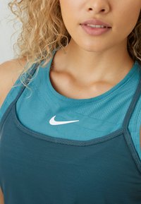Frau mit lockigem Haar, die geschichtete Sportoberteile in Teal und Blau von Nike trägt, mit leicht geöffneten Lippen, sichtbarem Schlüsselbein und Hals.