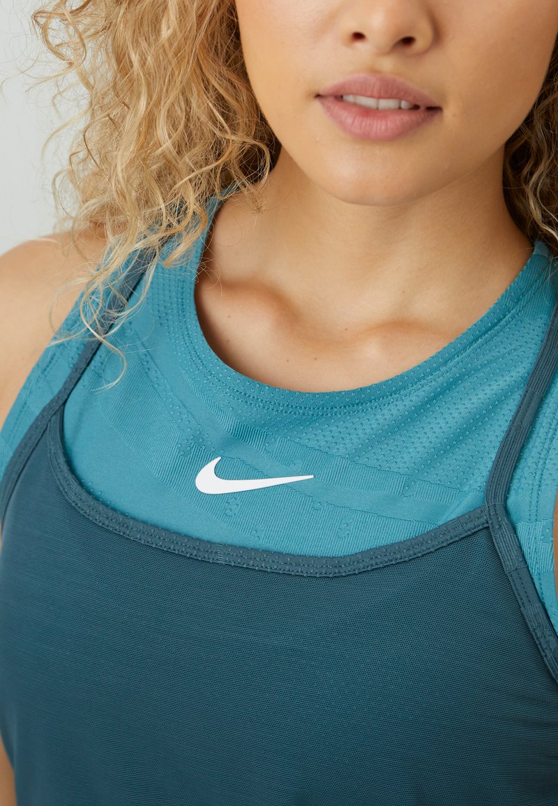 Frau mit lockigem Haar, die geschichtete Sportoberteile in Teal und Blau von Nike trägt, mit leicht geöffneten Lippen, sichtbarem Schlüsselbein und Hals.