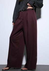 Pantalon à jambes larges en tissu bordeaux profond, avec des plis avant marqués, une silhouette ajustée et une ceinture noire, associé à des sandales noires.
