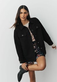 Femeie îmbrăcată într-o jachetă neagră cu nasturi, fustă cu model, șosete roșii și ghete negre până la gleznă, ajustând cămașa cu o mână.