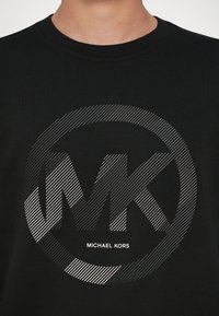 Μαύρη φούτερ μπλούζα με κυκλικό σχέδιο ασημί γκράντεντ και τα γράμματα "MK" καθώς και το κείμενο "MICHAEL KORS" από κάτω σε λευκό χρώμα.