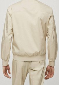 Beige letvægtsjakke med en struktureret ryg, elastiske manchetter og bund, rene linjer og minimalistisk design. Ingen synlige logoer eller mønstre.