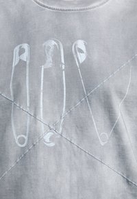 Siva dukserica s grafikom sigurnosnih igala u bijeloj boji, s vidljivim šavovima i mekim, teksturiranim pamukom.