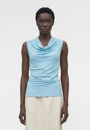 Γυναίκα με κοντά σγουρά μαλλιά που φοράει αμάνικο ανοιχτό μπλε τοπ με draping και μπεζ φούστα, στέκεται μπροστά σε ένα απλό λευκό φόντο.