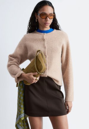 Vrouw met beige vest met knopen, donkerbruine rok, amberkleurige zonnebril, die een mosterdkleurige handtas vasthoudt met een groen sjaaltje met patroon.