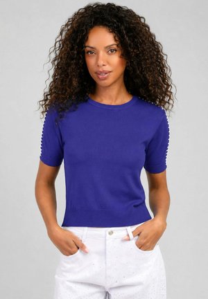 Femme aux cheveux bouclés portant un haut en maille violet à manches courtes et un pantalon blanc, debout les mains dans les poches devant un fond uni.