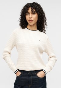 Kreminės spalvos megztinis iš megztinio su tekstūruotu rombų raštu, suapvalintu tamsiai mėlynos spalvos dryžuotu iškirptės kraštu ir ilgomis rankovėmis. Dėvimas su tamsiomis džinsinėmis kelnėmis.