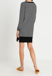 Vestido a rayas blanco y negro con mangas largas, escote redondo y dobladillo negro sólido. Modelo con calzado amarillo en un fondo neutro.
