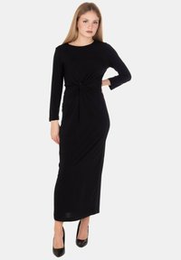 Abito nero a maniche lunghe con scollo rotondo, dettaglio twist in vita e silhouette aderente, indossato con tacchi neri. Tessuto liscio e lunghezza fino al pavimento.