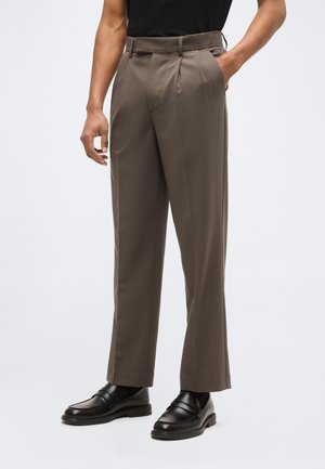 Mężczyzna noszący brązowe, plisowane spodnie z wysokim stanem, bocznymi kieszeniami i czarne skórzane mokasyny, stoi z jedną ręką w kieszeni.