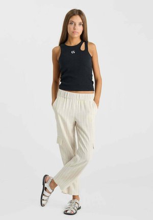 Jeune femme debout, les mains dans les poches, vêtue d'un haut noir sans manches avec découpe, d'un pantalon beige à fines rayures et de sandales argentées.