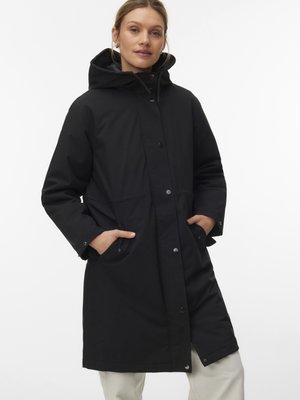 Femme portant un long manteau noir à capuche, les mains dans les poches avant, debout sur un fond blanc uni.