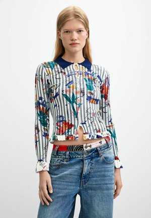 Junge Frau mit langen blonden Haaren, die ein gestreiftes, florales Langarmoberteil mit blauem Kragen und blaue Jeans vor einem schlichten Hintergrund trägt.