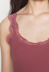 Primo piano di una donna che indossa un top senza maniche a costine in colore malva, con pizzo intorno al collo e alla spallina.