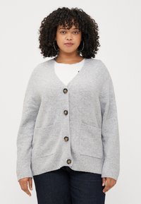 A woman with curly hair wears a gray cardigan over a white shirt, standing against a plain background. She has a neutral expression.