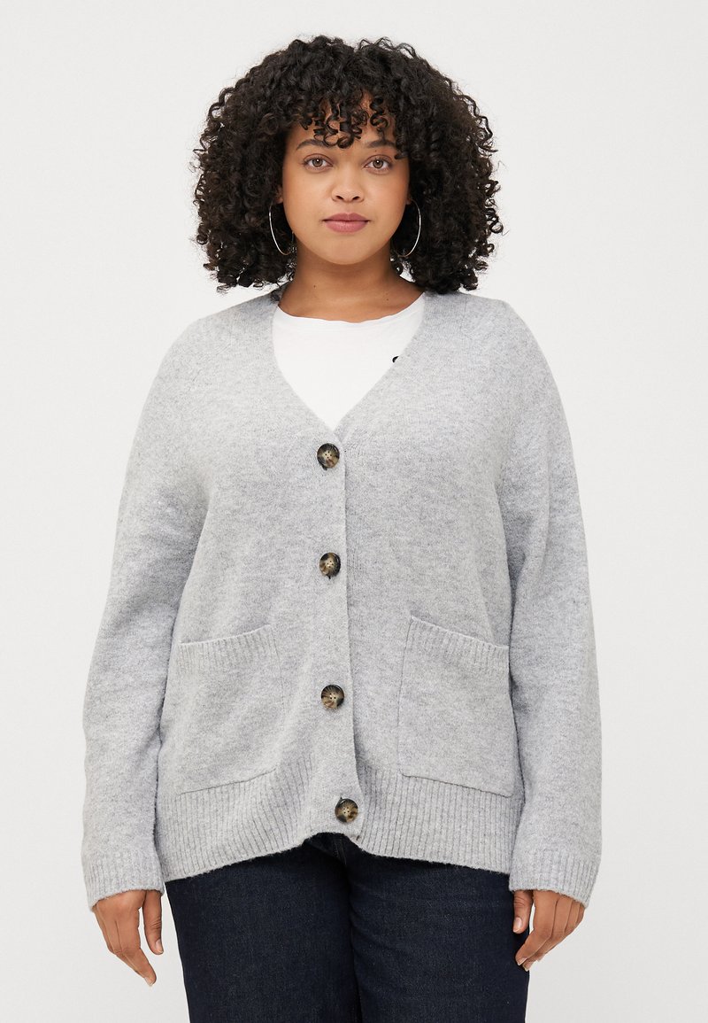 Une femme aux cheveux bouclés porte un cardigan gris par-dessus une chemise blanche, se tenant devant un fond uni. Elle a une expression neutre.
