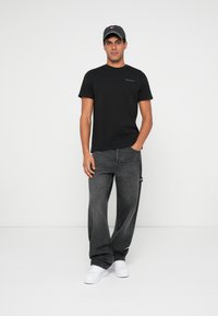 Camiseta negra de manga corta con un pequeño logotipo, combinada con unos vaqueros oscuros de corte holgado y zapatillas blancas. Un atuendo simple y casual.