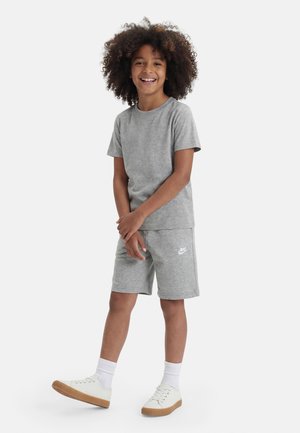 Uśmiechnięte dziecko o kręconych włosach, ubrane w szarą koszulkę Nike i krótkie spodenki, białe skarpetki oraz białe trampki, stojące na jednolitym tle.